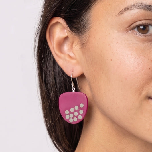 A close-up of a woman's ear wearing a pink guitar-pick-shaped dangle earring with white circular cutouts and a silver fishhook wire.