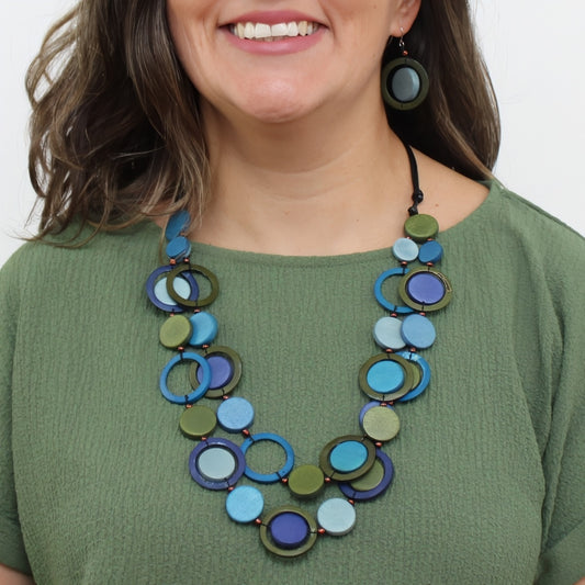 A woman wearing a colorful statement necklace made of overlapping circular discs in shades of blue, green, and purple, paired with matching round drop earrings, against a plain white background.
