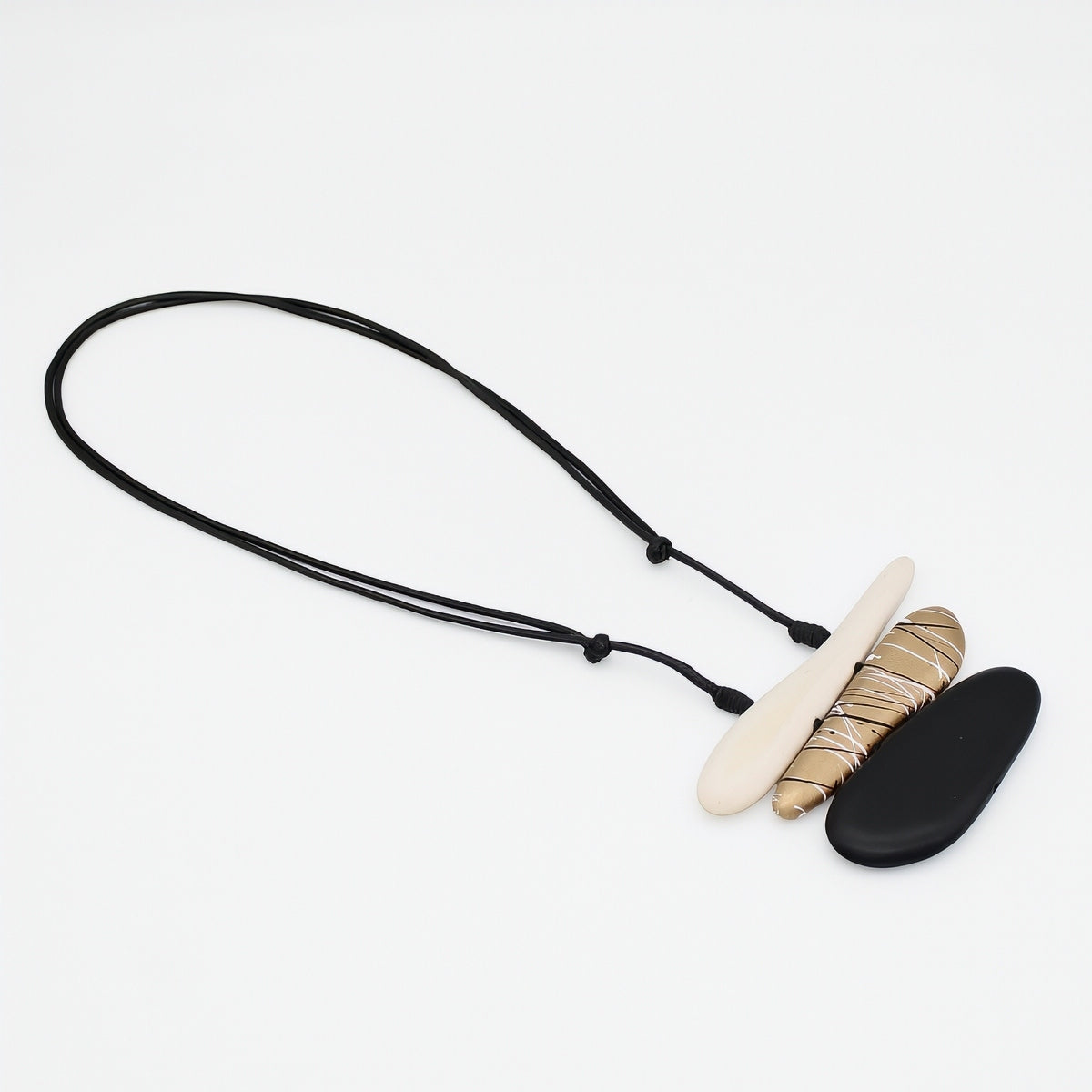 A modern pendant necklace featuring a black adjustable cord, a flat cream-colored stone, a textured gold and beige wrapped stone, and a smooth black stone, displayed on a clean white background.