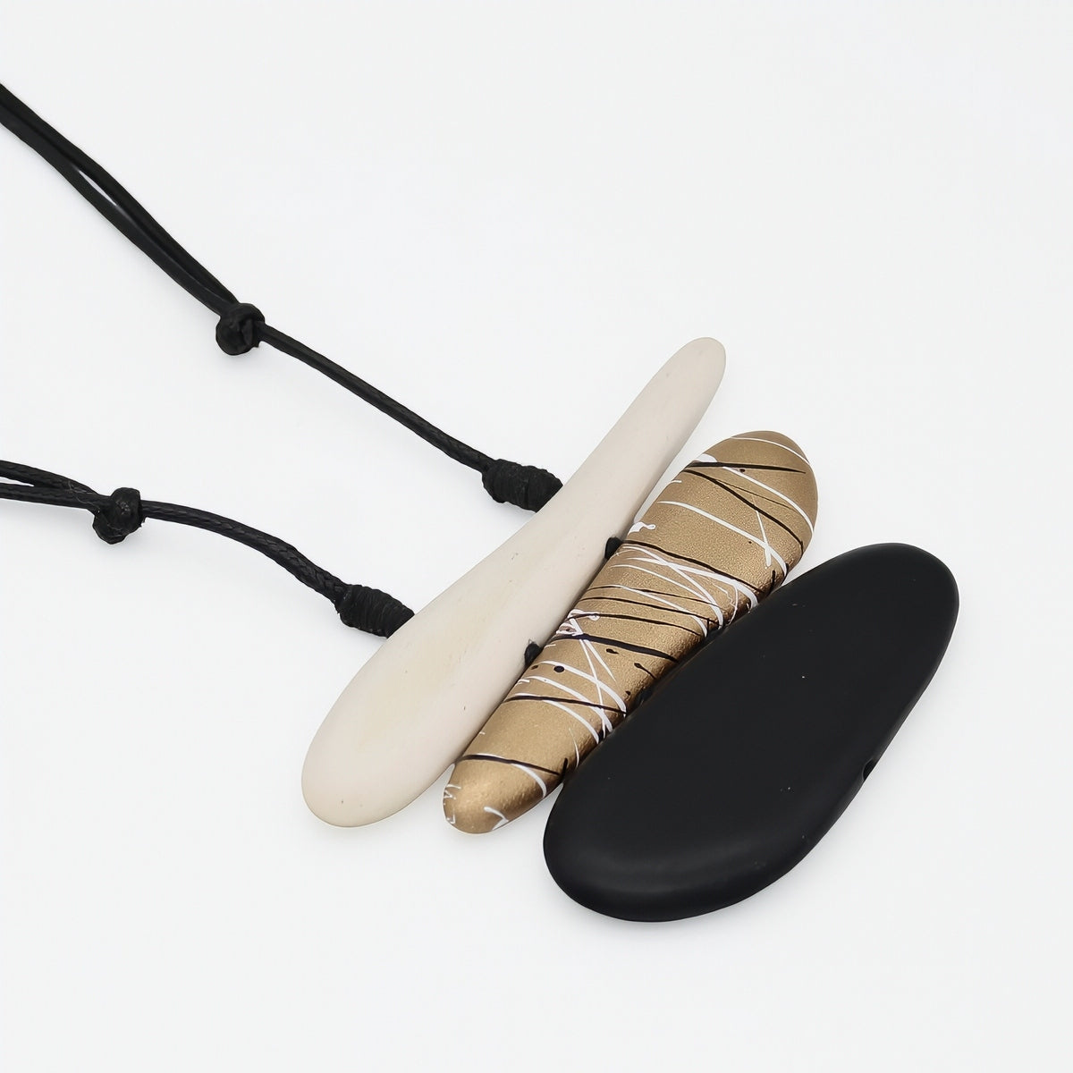 Three flat oval pendant necklaces on black cords, displayed on a clean white background. The necklaces feature matte black, matte cream, and a textured tan-and-white patterned pendant.