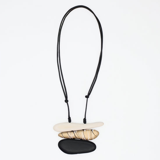 A minimalist necklace featuring three stacked natural stones in cream, tan, and black, suspended on a black adjustable cord against a plain white background.