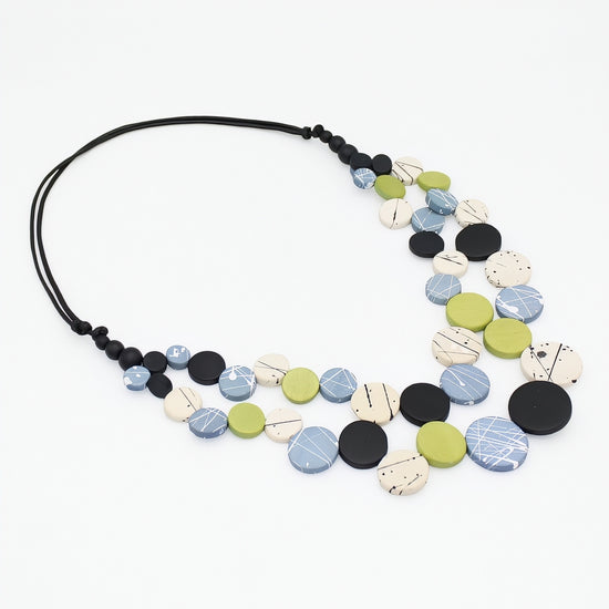 A multi-strand statement necklace featuring round, textured ceramic beads in black, light blue, olive green, and cream, suspended on a black cord, shown against a plain white background.