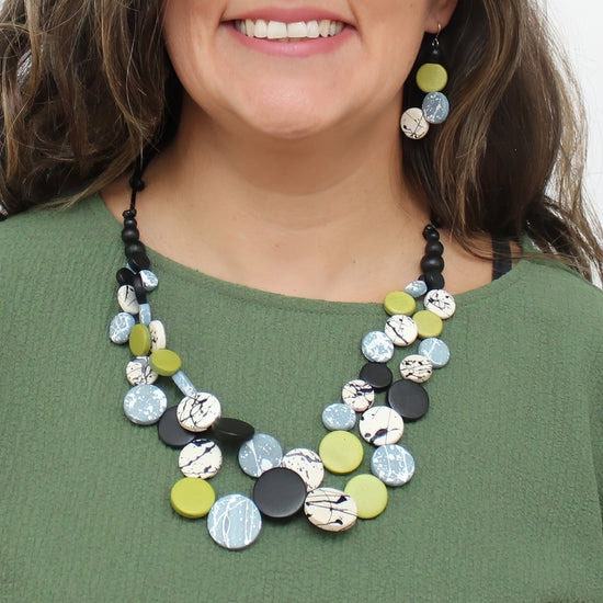 A close-up of a woman wearing a chunky multi-strand statement necklace and matching drop earrings. The jewelry features round disc elements in black, light blue, olive green, and white marbled patterns, worn over an olive green top.
