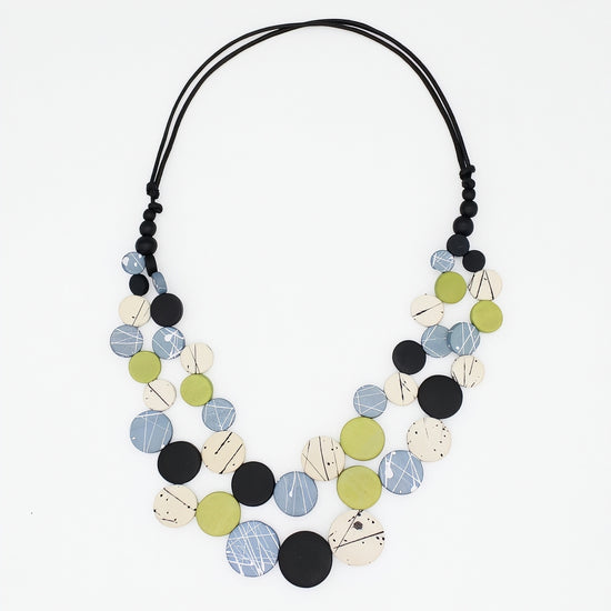 A multi-strand statement necklace featuring circular wooden discs in black, light blue, olive green, and cream with abstract line patterns, suspended on a black cord against a white background.