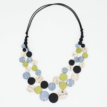 A multi-strand statement necklace featuring circular wooden discs in black, light blue, olive green, and cream with abstract line patterns, suspended on a black cord against a white background.