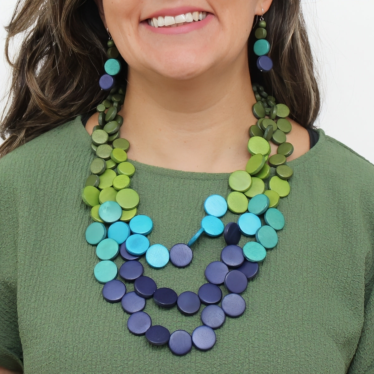 A close-up of a woman wearing a multi-strand statement necklace and matching drop earrings made of round wooden discs in shades of green, teal, and blue.