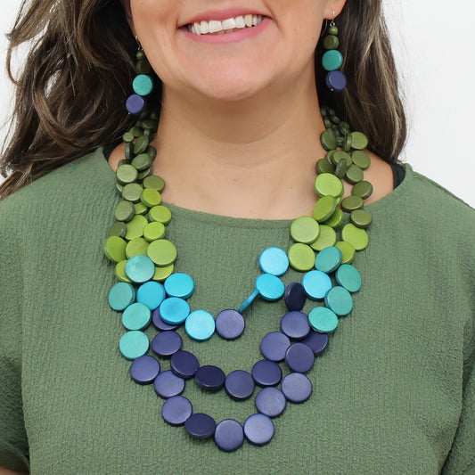 A close-up of a woman wearing a multi-strand statement necklace and matching drop earrings made of round wooden discs in shades of green, teal, and blue.