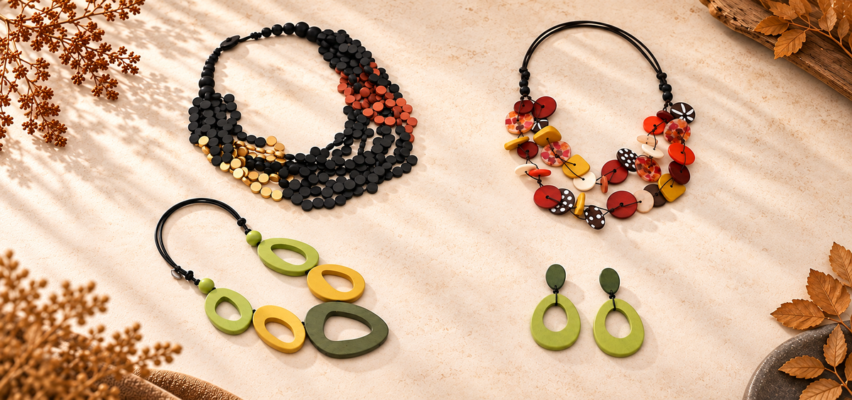 A styled flat-lay of handmade jewelry on a beige fabric background with dried botanicals. The set includes a multi-strand beaded necklace, a beaded necklace, a pair of green oval drop earrings, and a brown statement necklace