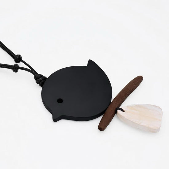 A minimalist pendant necklace featuring a matte black bird-shaped charm on a black cord, accompanied by a small brown wooden bead and a white mother-of-pearl teardrop charm, displayed on a clean white background.