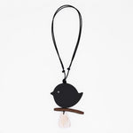 A minimalist black bird pendant necklace with a small wooden branch perch and a white tassel, suspended on a black cord against a plain white background.