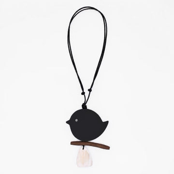 A minimalist black bird pendant necklace with a small wooden branch perch and a white tassel, suspended on a black cord against a plain white background.