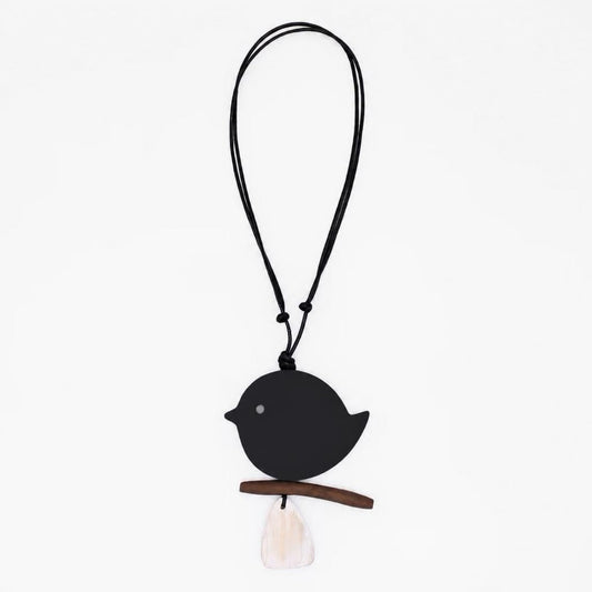 A minimalist black bird pendant necklace with a small wooden branch perch and a white tassel, suspended on a black cord against a plain white background.