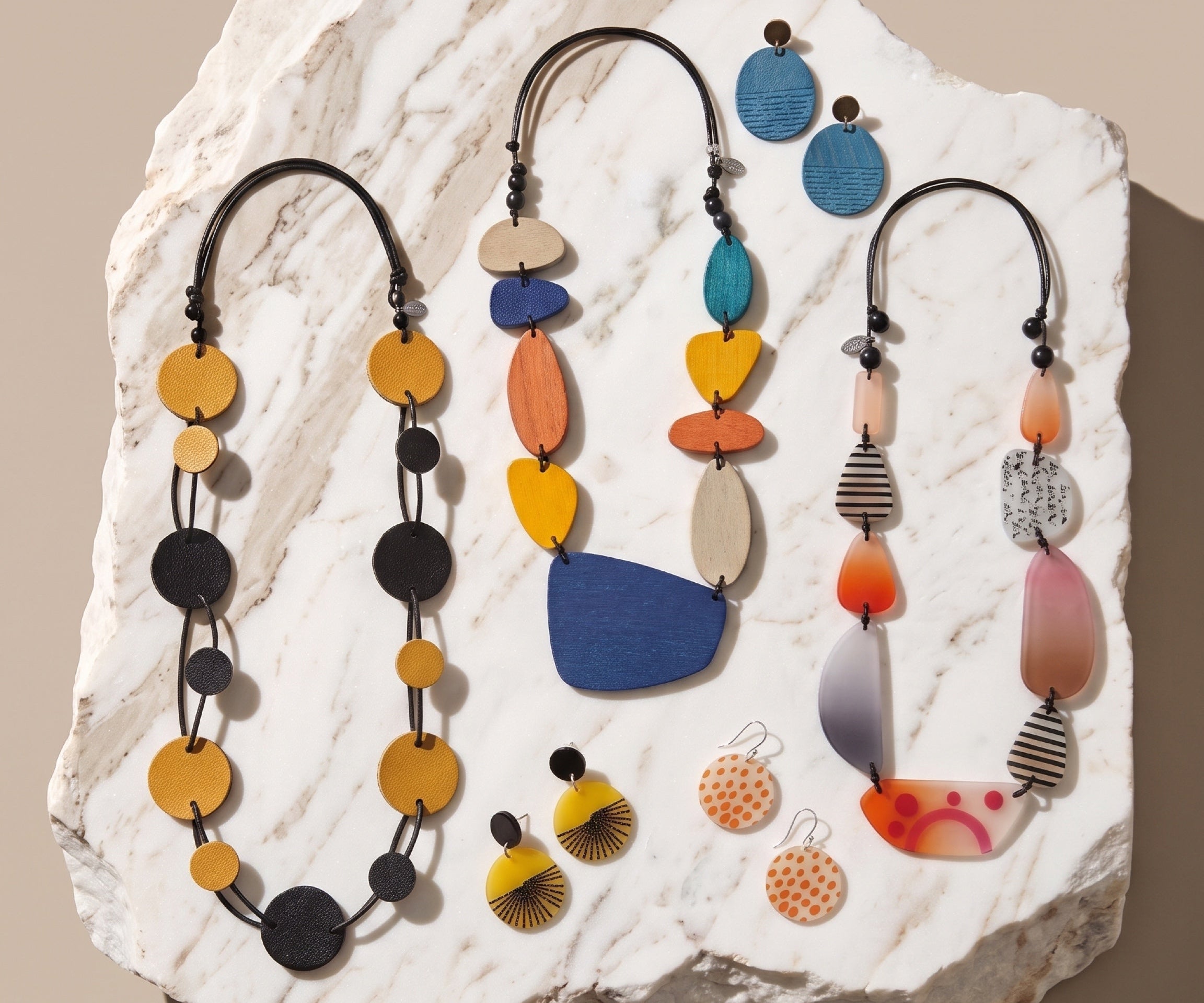 A styled flat-lay of colorful handmade jewelry, including three beaded necklaces and three matching earrings, arranged on a white marble slab against a solid beige background.