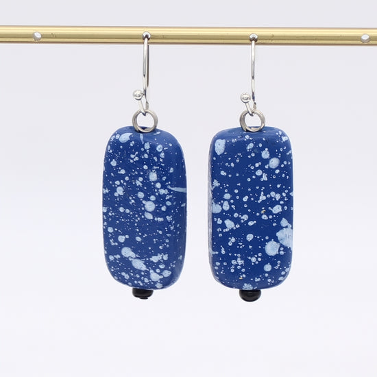 Blue speckled earrings hanging from a gold rod against a white background