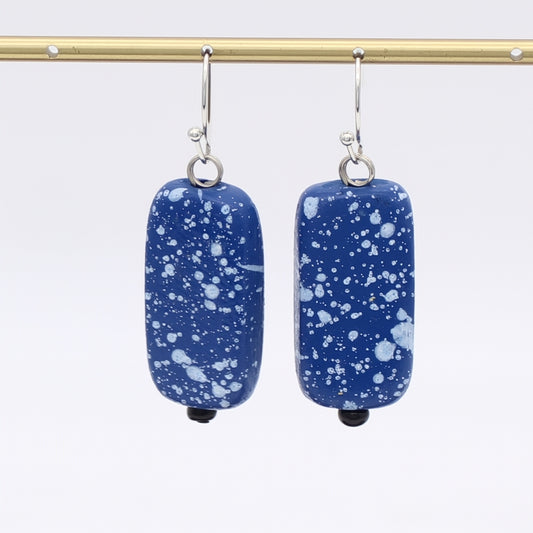 Blue speckled earrings hanging from a gold rod against a white background