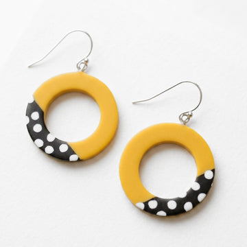 A pair of yellow circular drop earrings with a black and white polka dot pattern along the bottom half, hanging from silver fishhook wires on a white background.