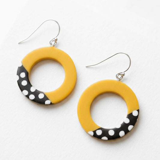 A pair of yellow circular drop earrings with a black and white polka dot pattern along the bottom half, hanging from silver fishhook wires on a white background.