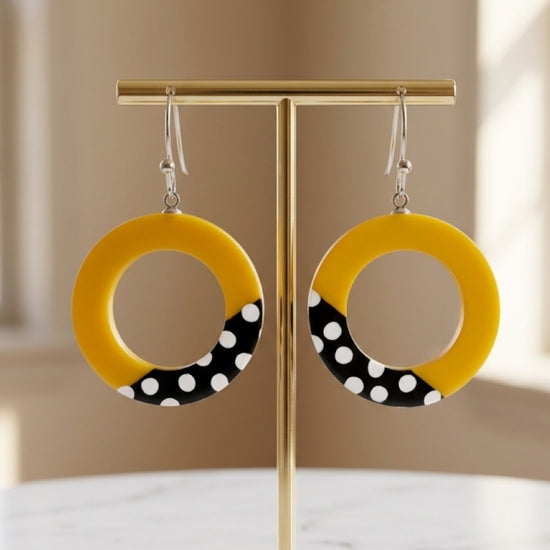 A pair of yellow circular drop earrings with a black and white polka dot section at the bottom, hanging from silver hooks on a gold T-stand against a softly blurred beige background.