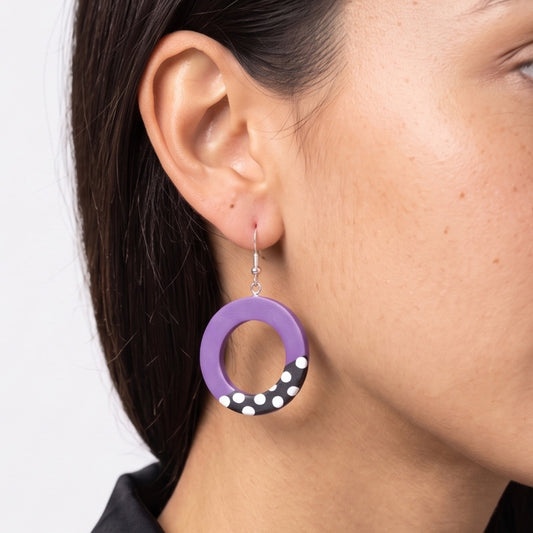 A close-up of a person's ear wearing a purple circular drop earring with a black and white polka dot accent at the bottom and a silver fishhook wire.