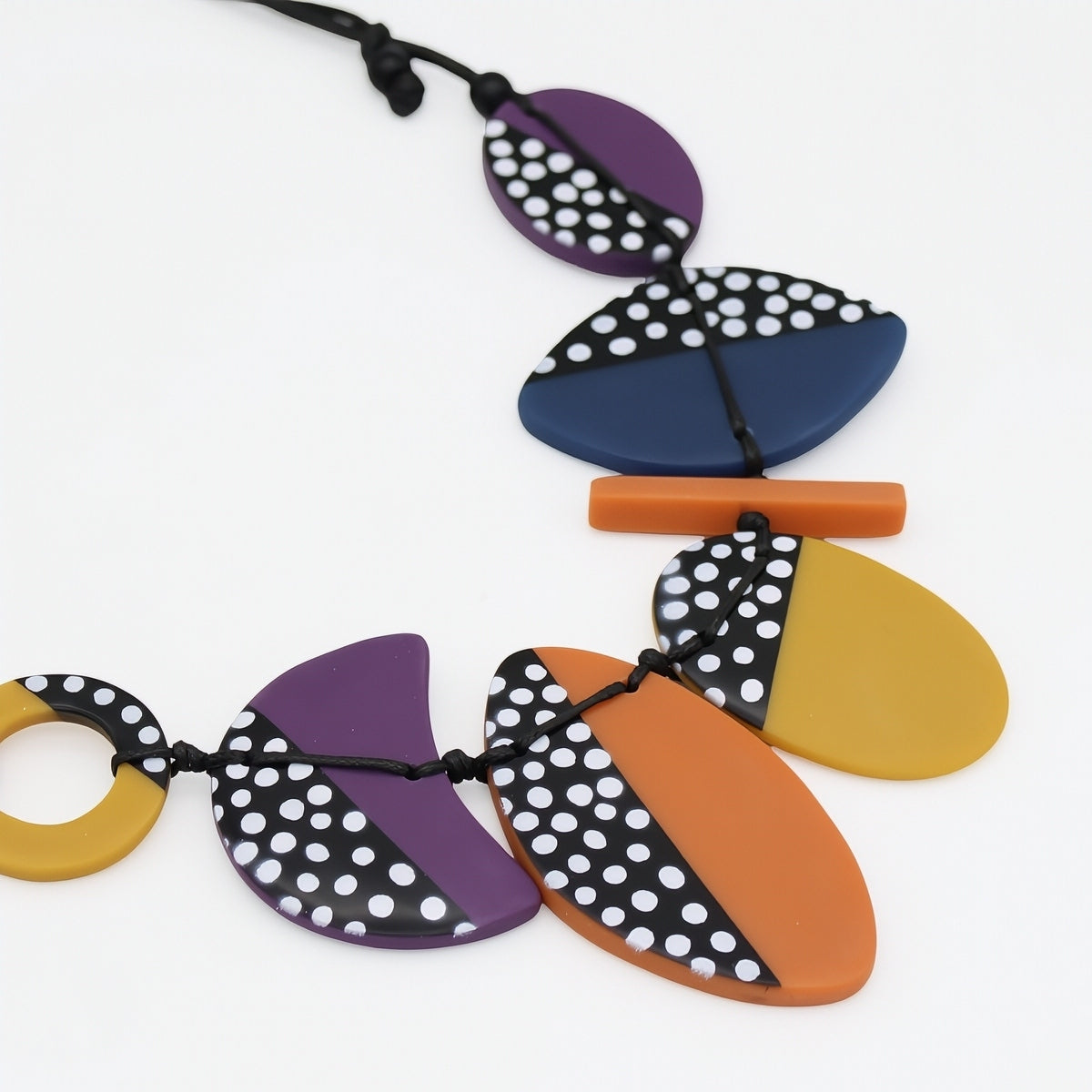 A colorful geometric pendant necklace on a black cord, featuring semi-circular and teardrop-shaped beads in purple, navy blue, orange, and mustard yellow, with some beads decorated in a black and white polka dot pattern.