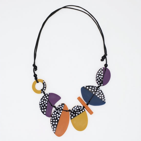 A multicolor geometric statement necklace with wooden-like disc and oval pieces in purple, navy, orange, and yellow, some with black and white polka dots, on a black cord, shown against a plain white background.