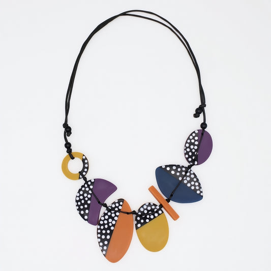 A multicolor geometric statement necklace with wooden-like disc and oval pieces in purple, navy, orange, and yellow, some with black and white polka dots, on a black cord, shown against a plain white background.