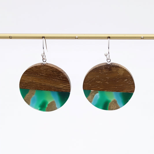 A pair of round drop earrings with a natural wood top half and a green and blue marbled bottom half, hanging from silver hooks on a gold rod against a white background.