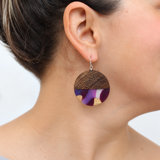 A close-up of a person's ear wearing a round drop earring with a brown wood-grain top half and a purple, white, and gold abstract patterned bottom half.