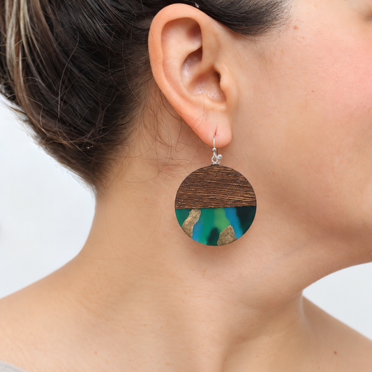 A close-up of a person's ear and neck wearing a round drop earring. The earring has a dark brown wood-grain top half and a bottom half featuring iridescent green, blue, and gold inlay.