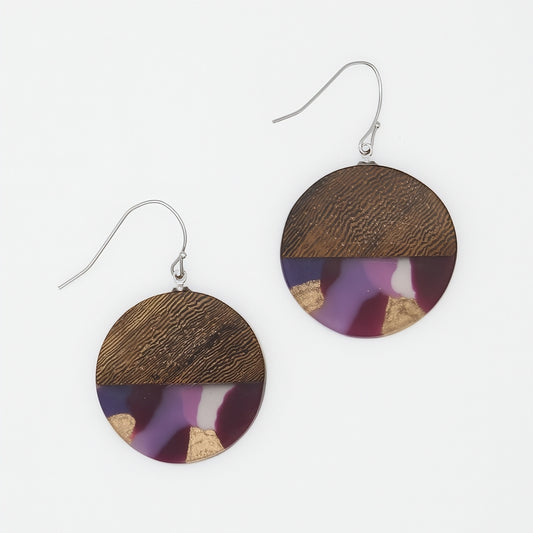 A pair of round drop earrings with a split design: the top half is natural brown wood grain, and the bottom half is a purple and white marbled resin with small gold leaf accents. They feature silver-tone fishhook wires, shown on a clean white background.