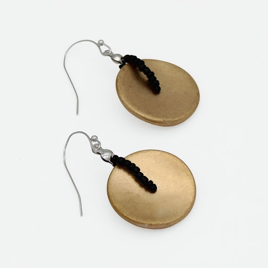 A pair of handmade dangle earrings featuring textured, oval-shaped gold-toned disc pendants with a small black beaded accent and silver fishhook wires, displayed on a clean white background.