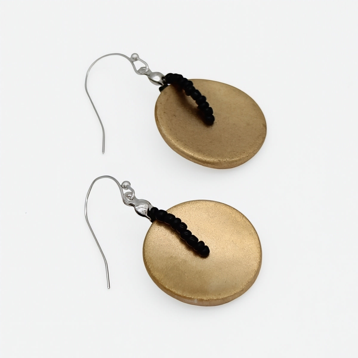 A pair of handmade dangle earrings featuring textured, oval-shaped gold-toned disc pendants with a small black beaded accent and silver fishhook wires, displayed on a clean white background.