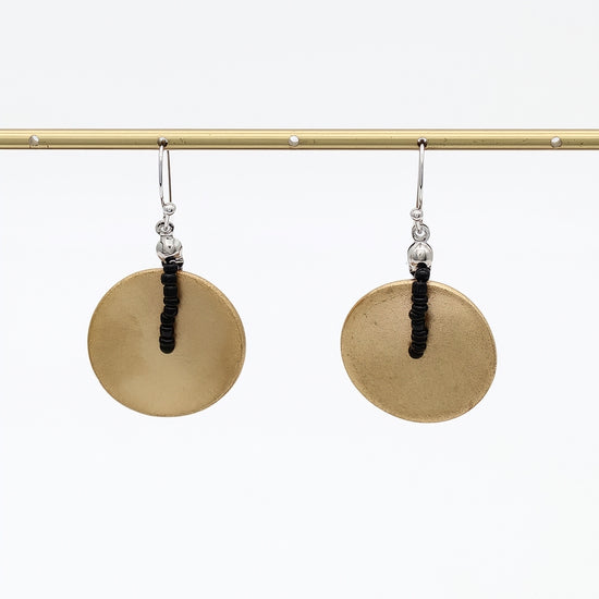 A pair of handmade dangle earrings with large, flat circular gold-toned discs and a central black beaded vertical accent, hanging from silver-toned hooks on a gold display bar against a white background.