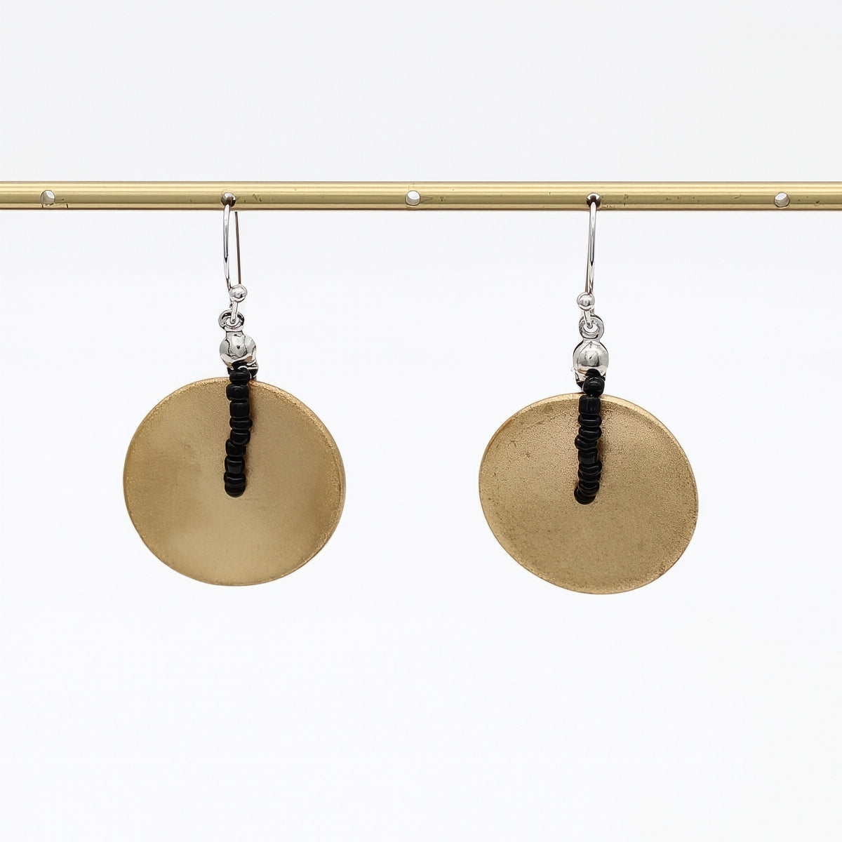 A pair of handmade dangle earrings with large, flat circular gold-toned discs and a central black beaded vertical accent, hanging from silver-toned hooks on a gold display bar against a white background.