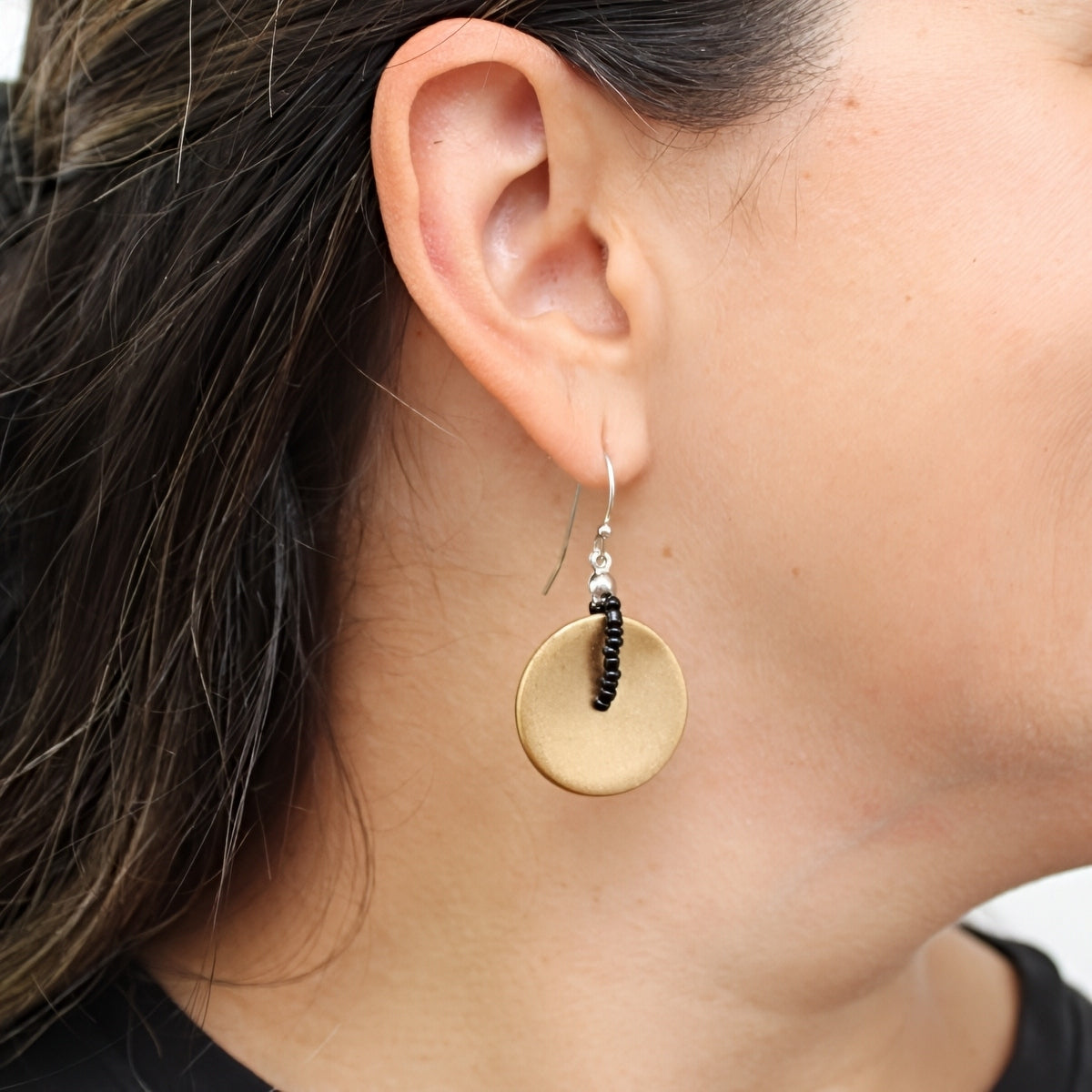A close-up of a person's ear wearing a dangling earring. The earring features a flat, circular gold-toned disc with a small black beaded vertical accent and silver hooks.