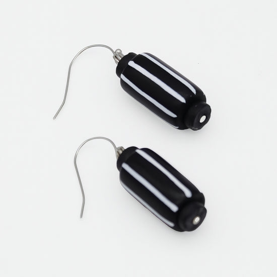A pair of drop earrings featuring glossy black cylindrical beads with three horizontal white stripes, suspended from silver fishhook wires on a plain white background.