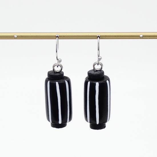 A pair of black cylindrical drop earrings with vertical white stripes, hanging from silver-toned hooks from a gold horizontal bar against a white background.