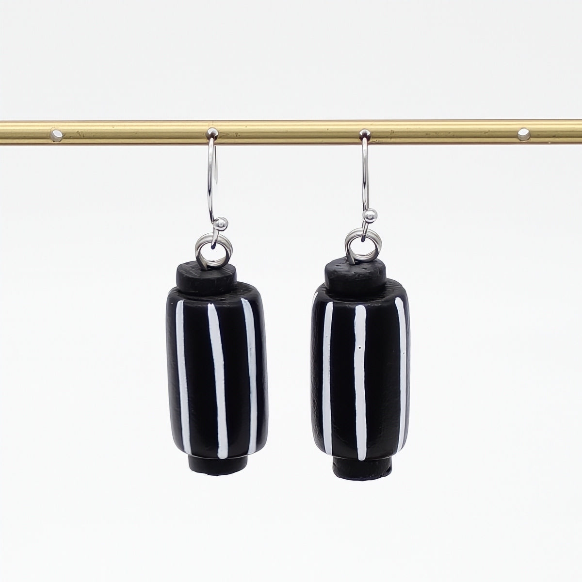 A pair of black cylindrical drop earrings with vertical white stripes, hanging from silver-toned hooks from a gold horizontal bar against a white background.