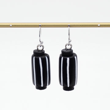 A pair of black cylindrical drop earrings with vertical white stripes, hanging from silver-toned hooks from a gold horizontal bar against a white background.