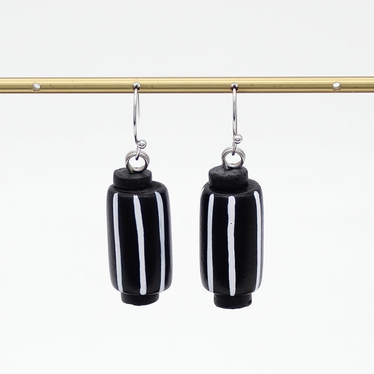 A pair of black cylindrical drop earrings with vertical white stripes, hanging from silver-toned hooks from a gold horizontal bar against a white background.