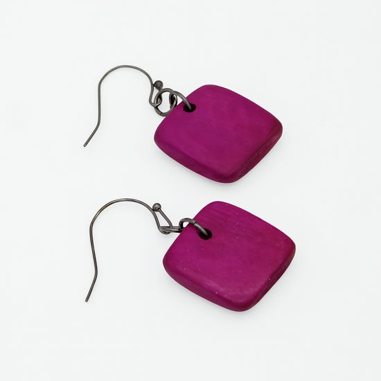 A pair of square-shaped dangle earrings made of textured magenta purple wood, attached to dark metal fishhook wires, shown on a plain white background.