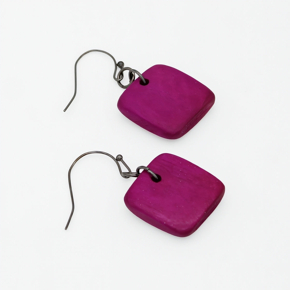 A pair of square-shaped dangle earrings made of textured magenta purple wood, attached to dark metal fishhook wires, shown on a plain white background.
