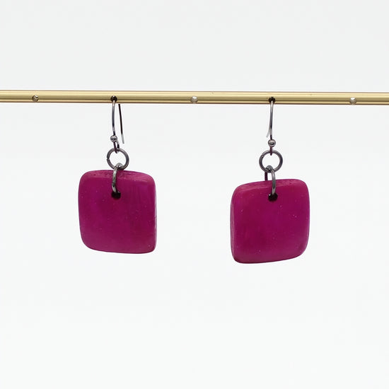 A pair of square-shaped magenta pink drop earrings with silver-toned hooks, hanging from a gold-colored horizontal bar against a plain white background.