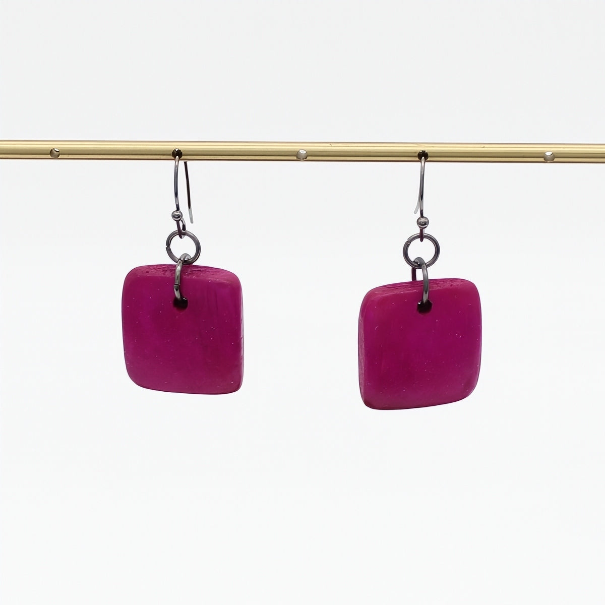 A pair of square-shaped magenta pink drop earrings with silver-toned hooks, hanging from a gold-colored horizontal bar against a plain white background.