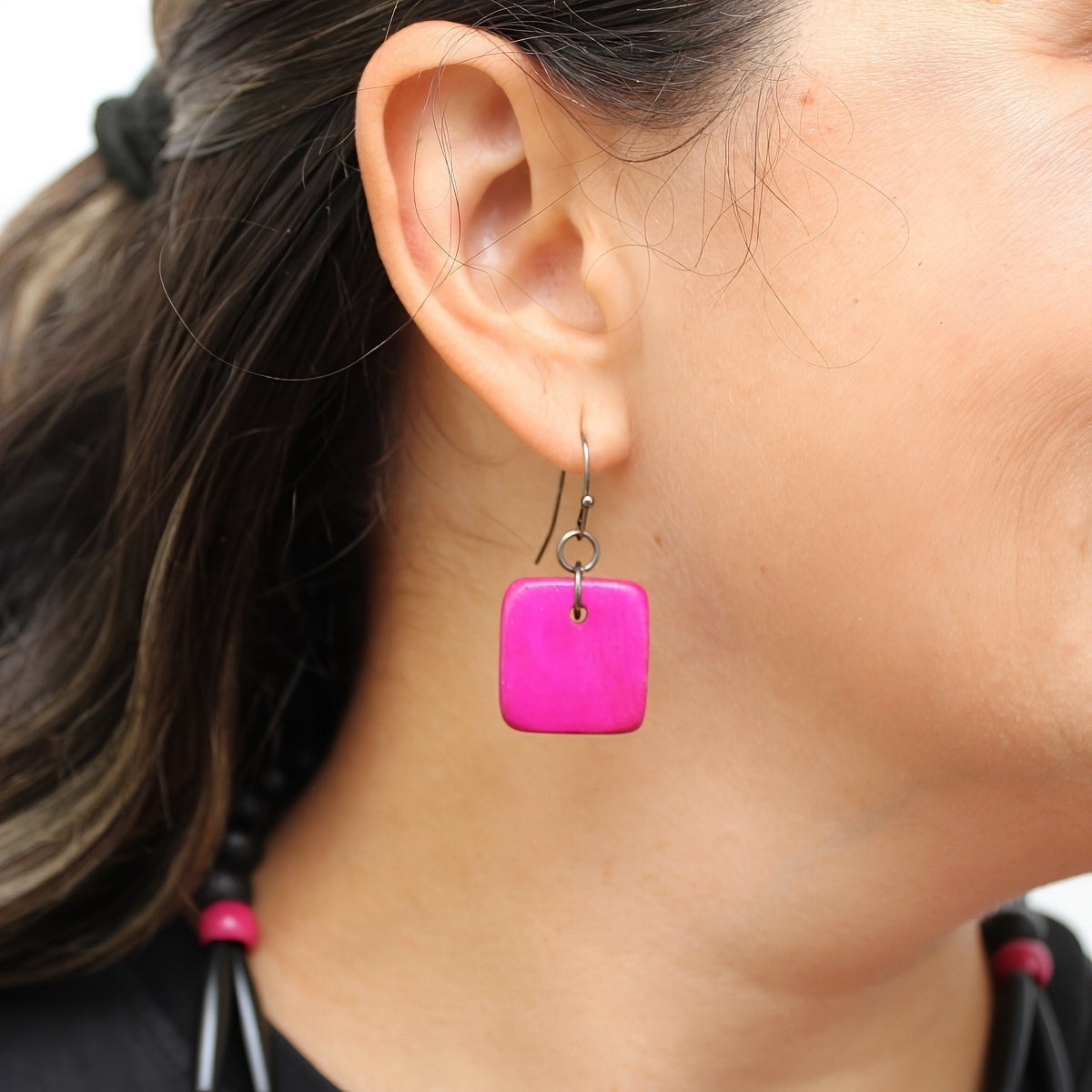 A close-up of a person's ear wearing a bright pink square drop earring with a silver fishhook wire.