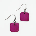 A pair of square-shaped drop earrings with a textured magenta-purple wooden disc and a dark metal fishhook ear wire, shown on a plain white background.