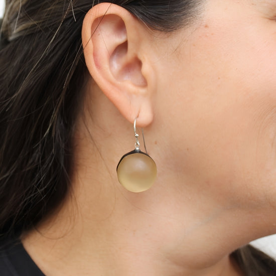 A close-up of a person's ear wearing a single drop earring with a round, matte, translucent beige/gold stone in a silver-toned setting.