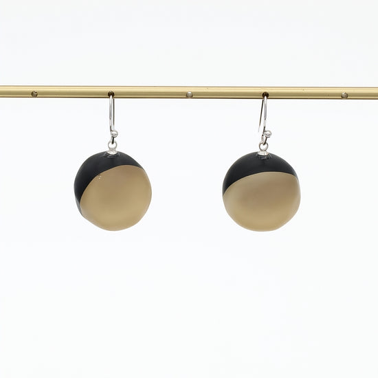 A pair of modern dangle earrings with a split black and golf design, hanging from silver hooks on a gold horizontal bar against a white background.