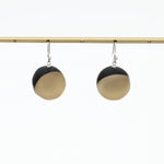 A pair of modern dangle earrings with a split black and golf design, hanging from silver hooks on a gold horizontal bar against a white background.