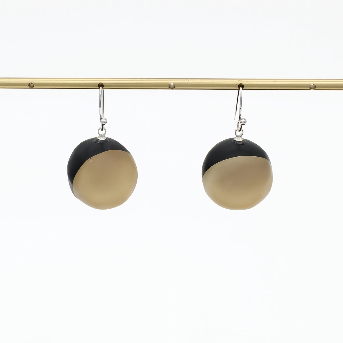 A pair of modern dangle earrings with a split black and golf design, hanging from silver hooks on a gold horizontal bar against a white background.
